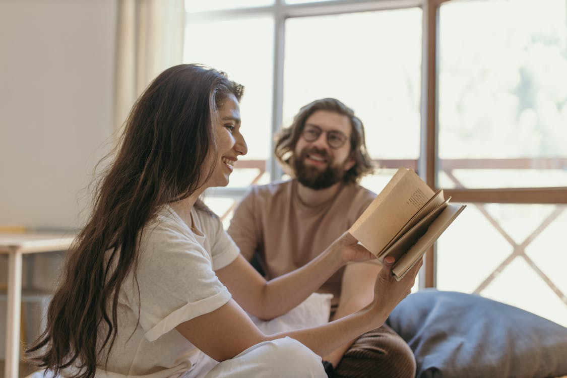 Happy couple in bed laughing together as the woman reads a book, symbolizing restored intimacy and connection through hormone therapy.